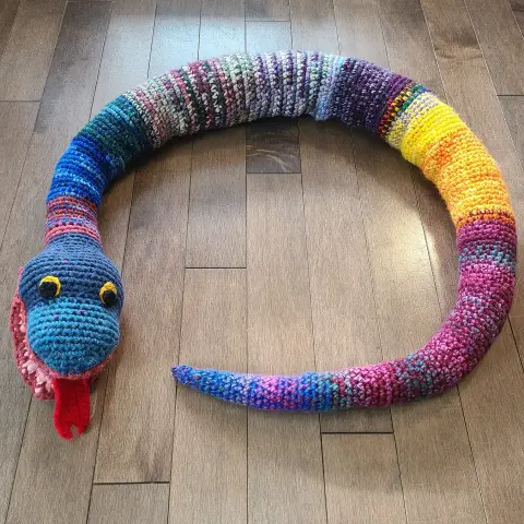 An overhead picture of scrappy snake. It has stripes and speckles in many colours. It has round eyes with black pupils and yellow irises. It has a red forked tongue sticking out of a pink mouth and red lips. It is curled almost into a circle. The body gets wider in the middle, then narrows again at the tail to a point.