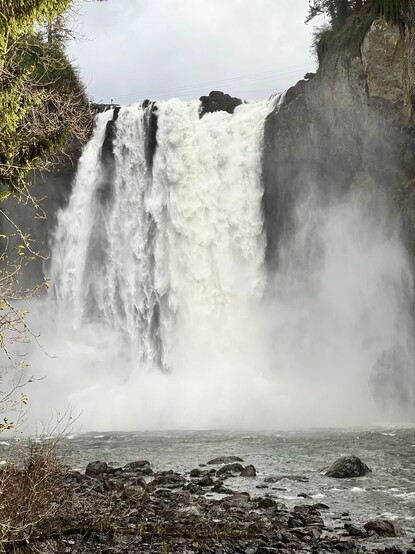 View of the bottom of Snoqualmie Falls with water spraying into the air and rocky shoreline in the foreground.