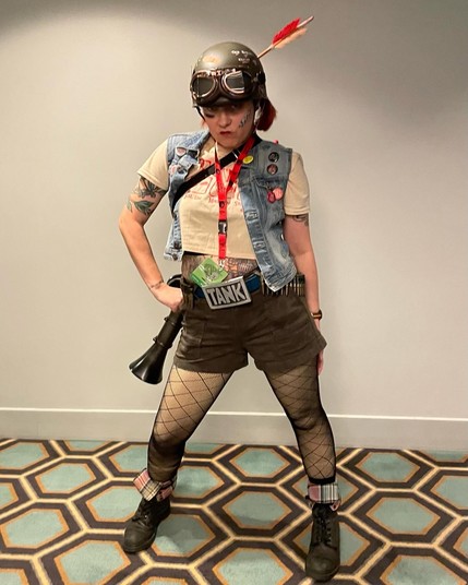A cosplayer (me!) in a hallway with hexagonal carpeting. They are wearing boots, shorts, a crop top, a denim vest, fishnet tights and an army helmet with goodies affixed and an arrow sticking out, carrying a bazooka strapped across their back. 