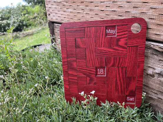 Wooden puzzle calendar showing date Sat May 18th against wooden crate among wildflowers.