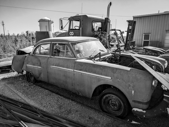 Black and white photograph. An abandoned old car, vintage early 1950s, sits in a boneyard of other abandoned things.