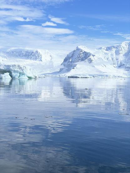 Snow covered mountains rise out of the ocean. An iceberg breaks through the water in front of the mountains. The sky is blue with minimal wispy white clouds. 